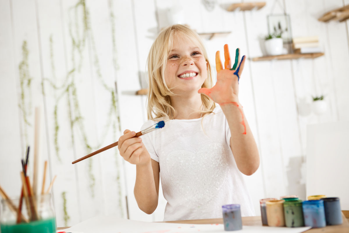 happy-playful-cute-freckled-blonde-girl-dressed-white-holding-brush-one-hand-showing-another-hand-which-she-messed-up-with-paint